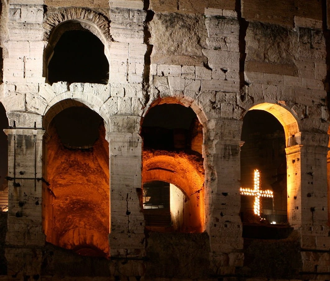 Stone arches of the Colosseum at night, lit from within by warm light and a large, illuminated cross.