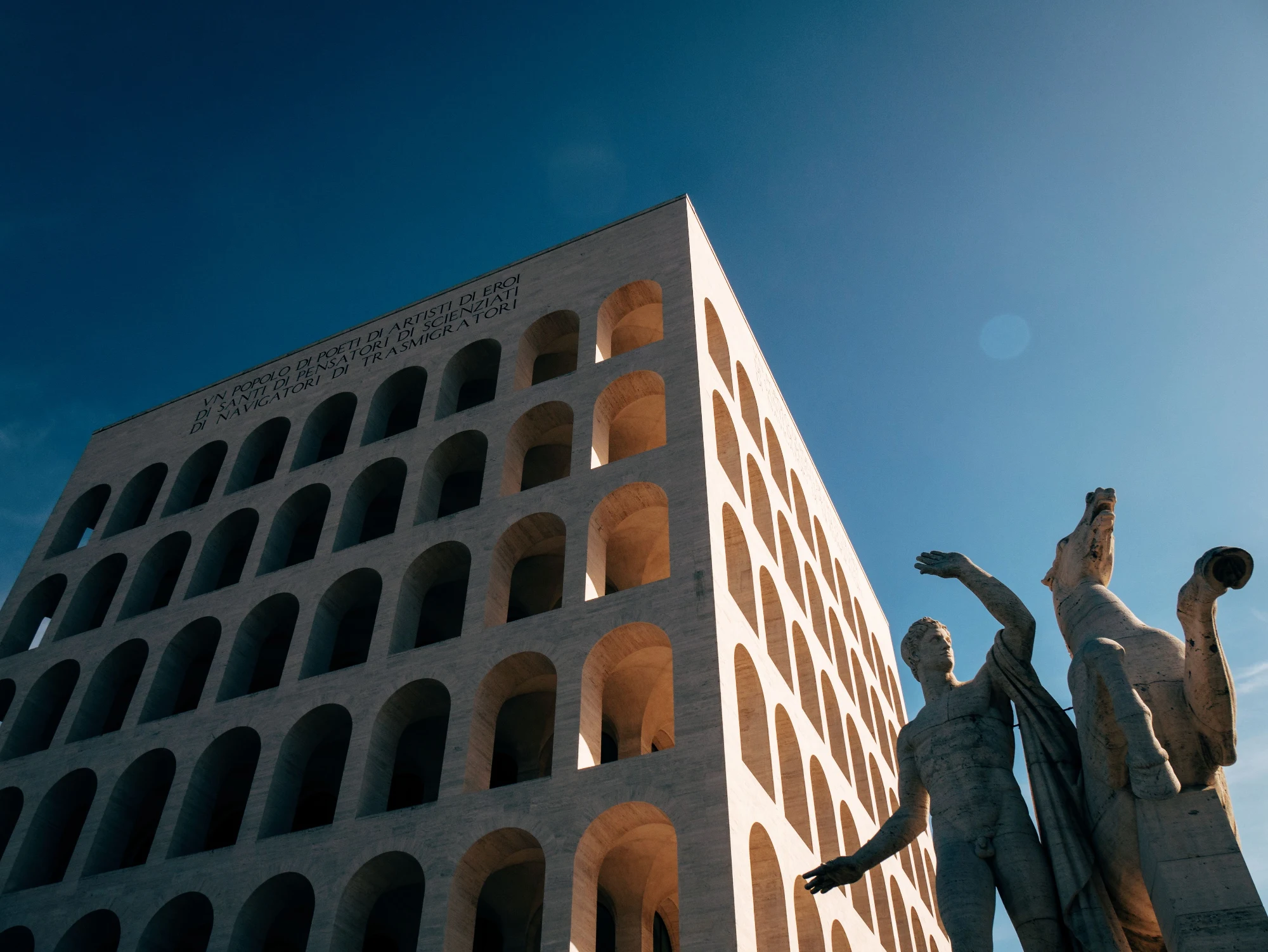 The Palazzo della Civiltà Italiana in Rome's EUR district, viewed from below against a clear blue sky, with a statue in th...
