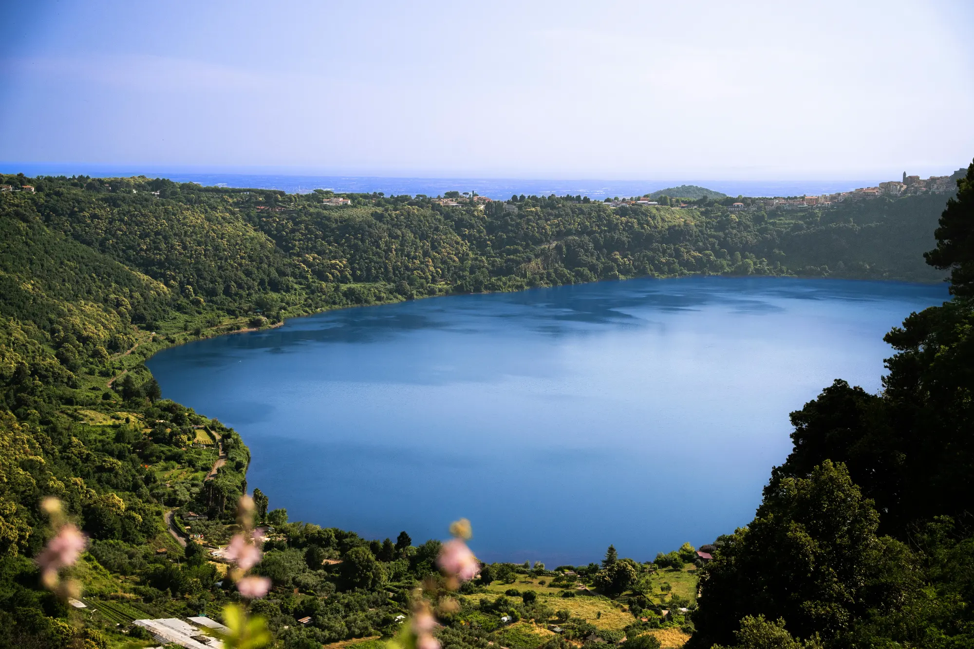 A wide view of the deep blue Lago Albano, a volcanic lake surrounded by lush green hills and small towns on its rim.