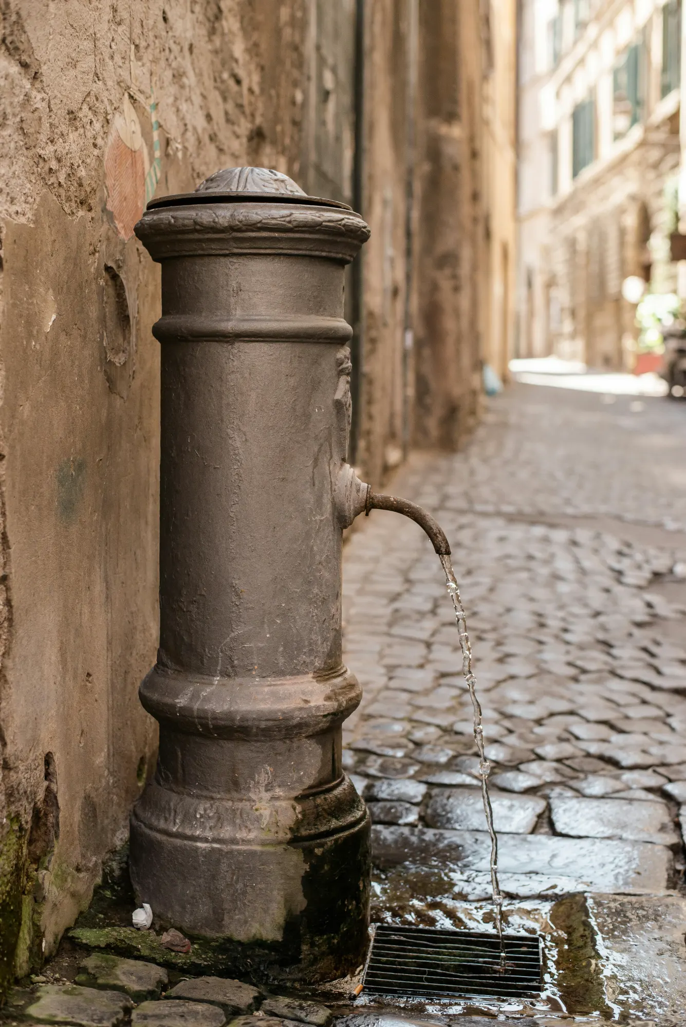 Traditional cast-iron nasone drinking fountain with flowing water on a cobblestone street in Rome.