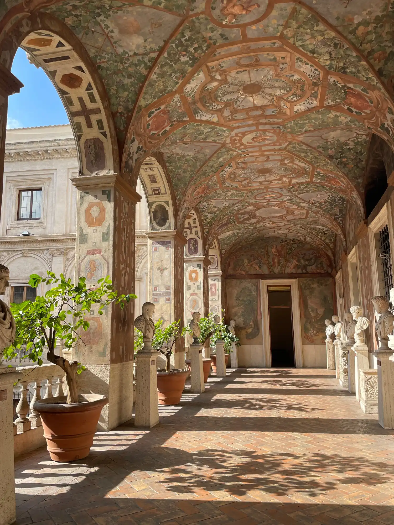 A sunlit palace loggia with a frescoed ceiling, arched colonnade, and a row of classical busts on pedestals.
