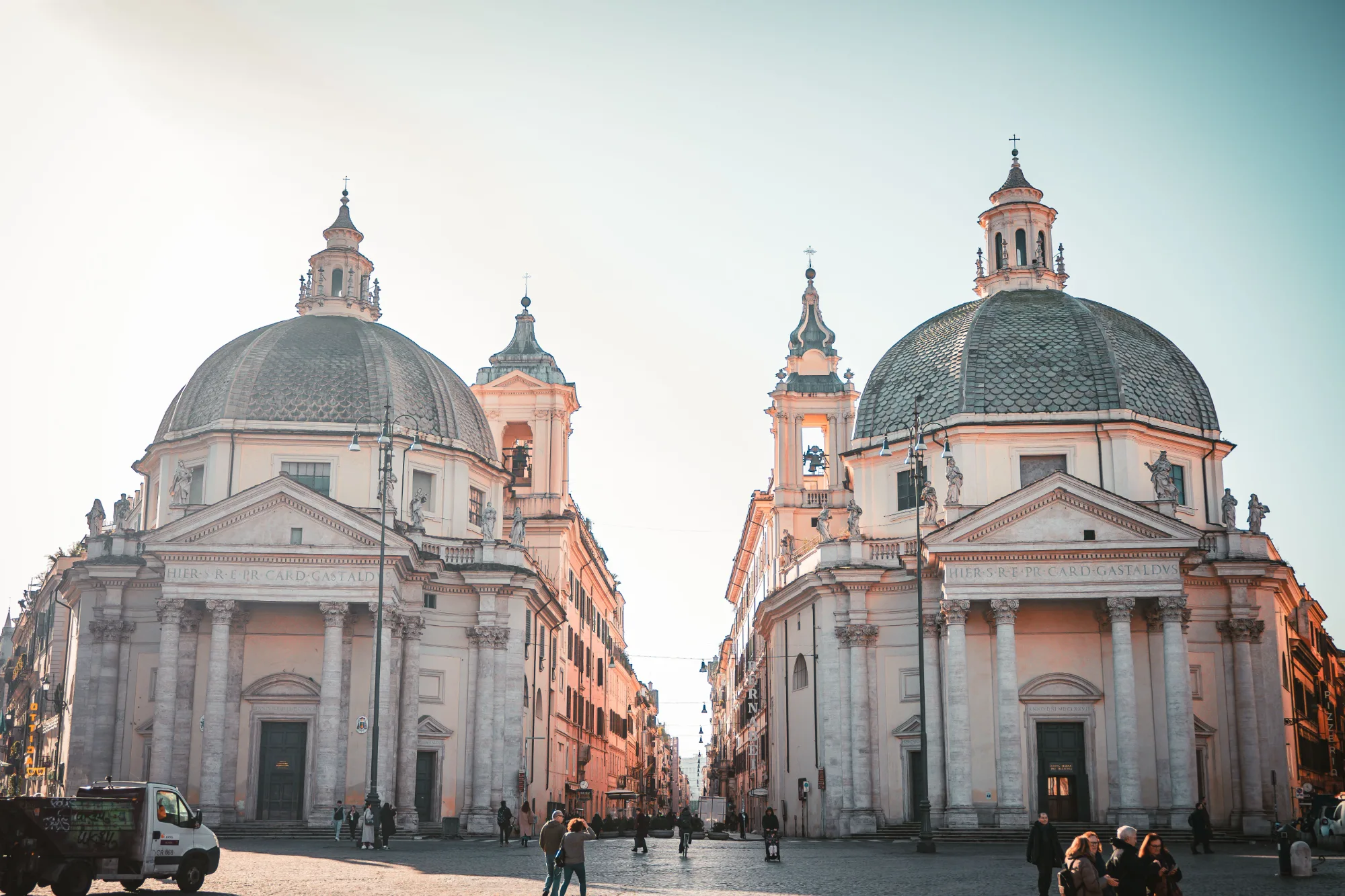 The twin churches of Piazza del Popolo in Rome, flanking a street at sunrise.