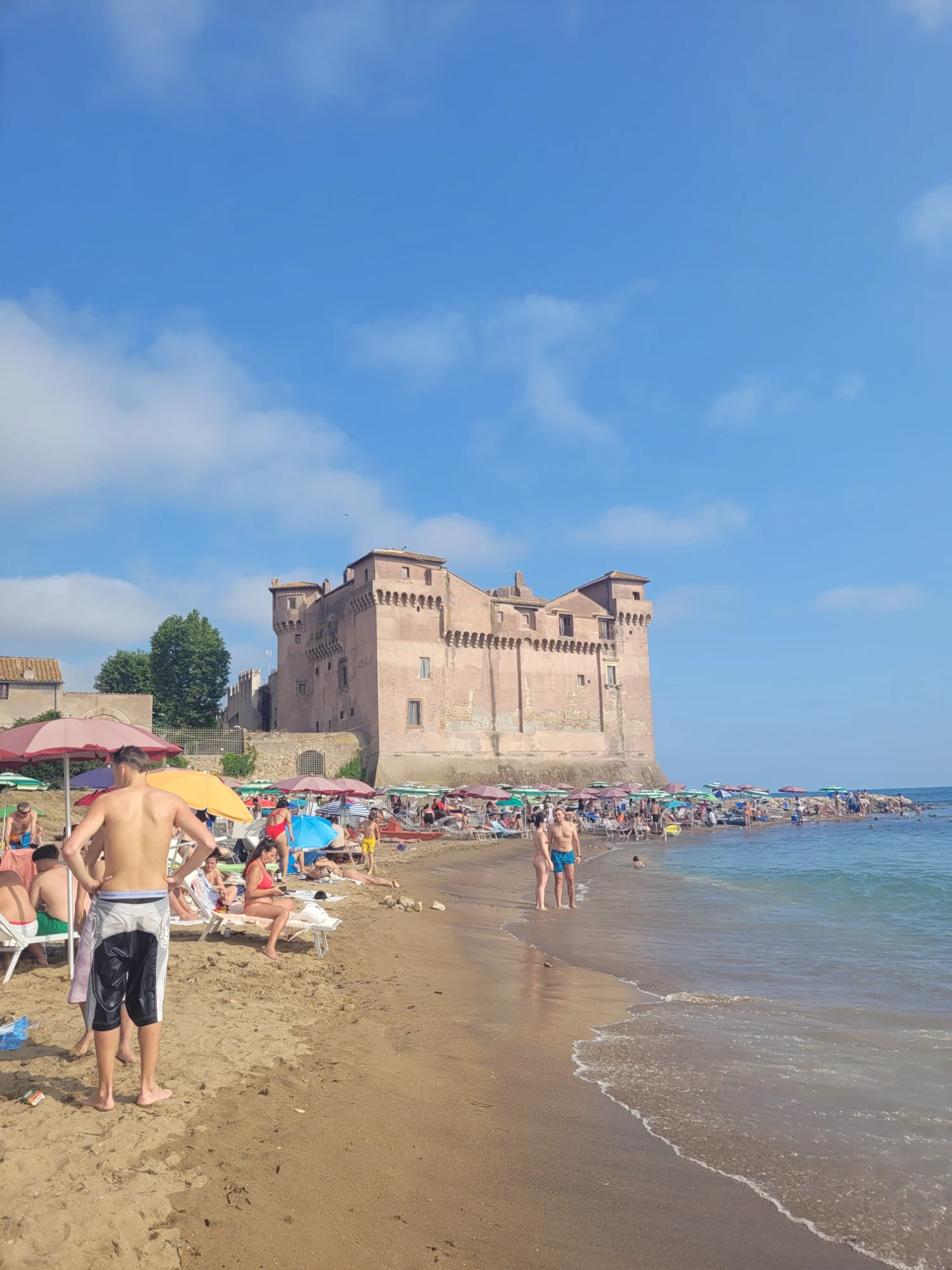 A busy sandy beach next to the medieval Castle of Santa Severa under a blue sky.