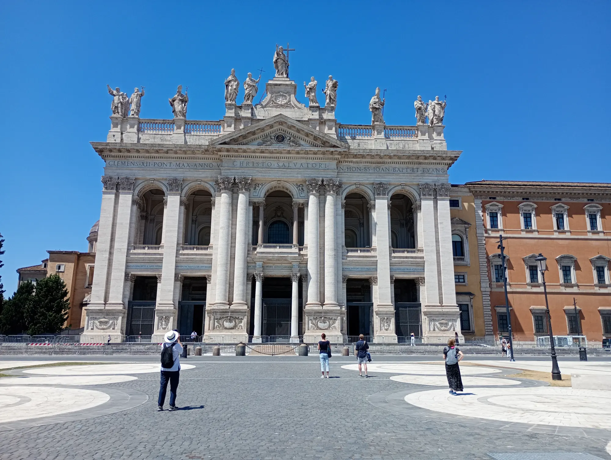 The grand facade of the Archbasilica of St. John Lateran in Rome under a clear blue sky, with a few tourists in the square.
