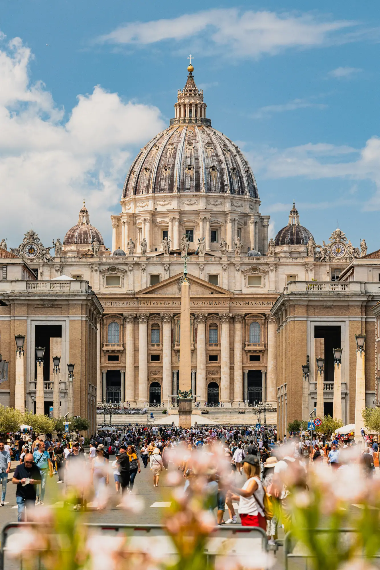 St. Peter's Basilica dome and facade under a blue sky, with an obelisk and crowds of people in St. Peter's Square.