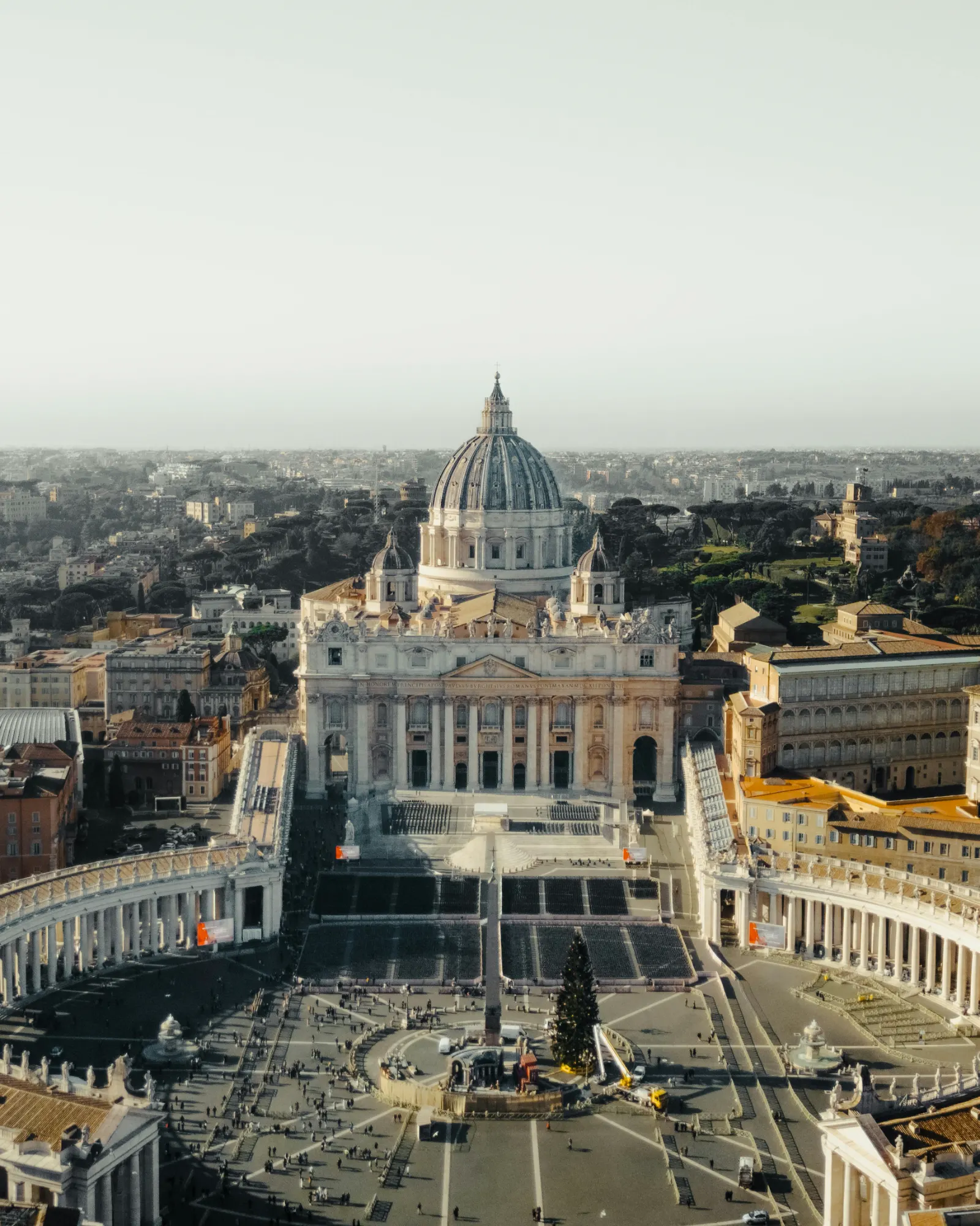 Aerial view of St. Peter's Basilica and its square, crowded with people, with the city of Rome spreading in the distance.