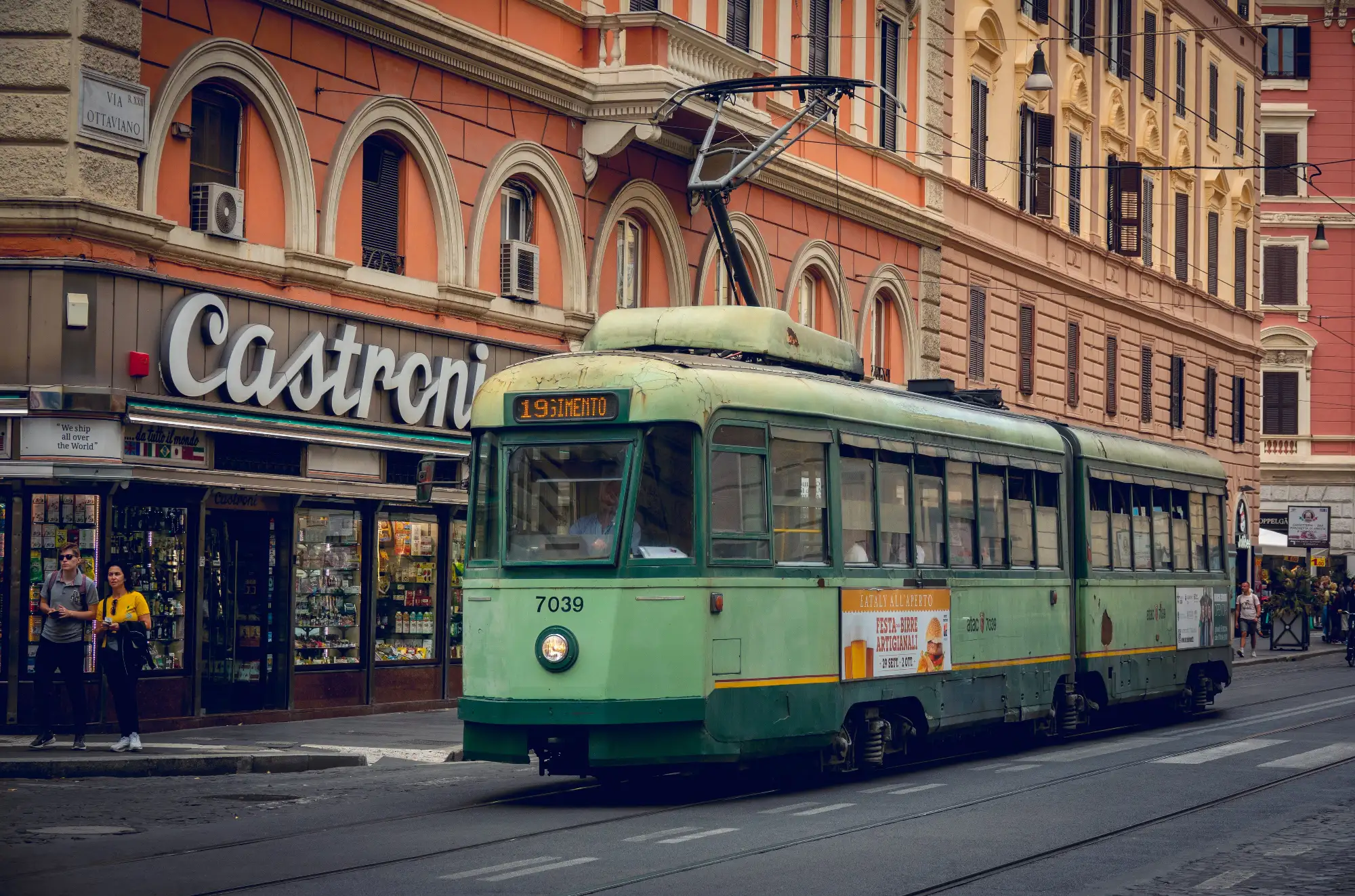 A vintage green tram on a street in Rome, with a 'Castroni' shop and orange buildings in the background.
