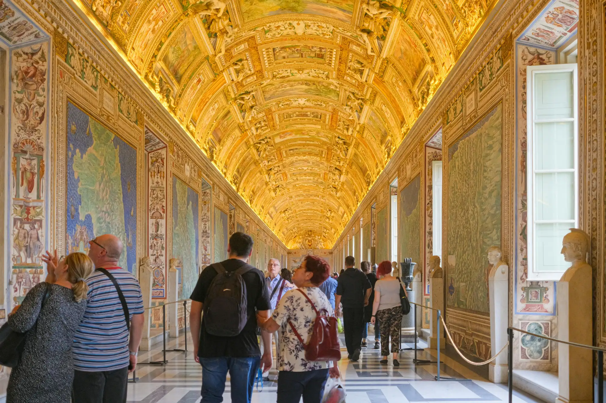 Tourists inside the Gallery of Maps in the Vatican, with its ornate, gilded barrel-vault ceiling and map-covered walls.