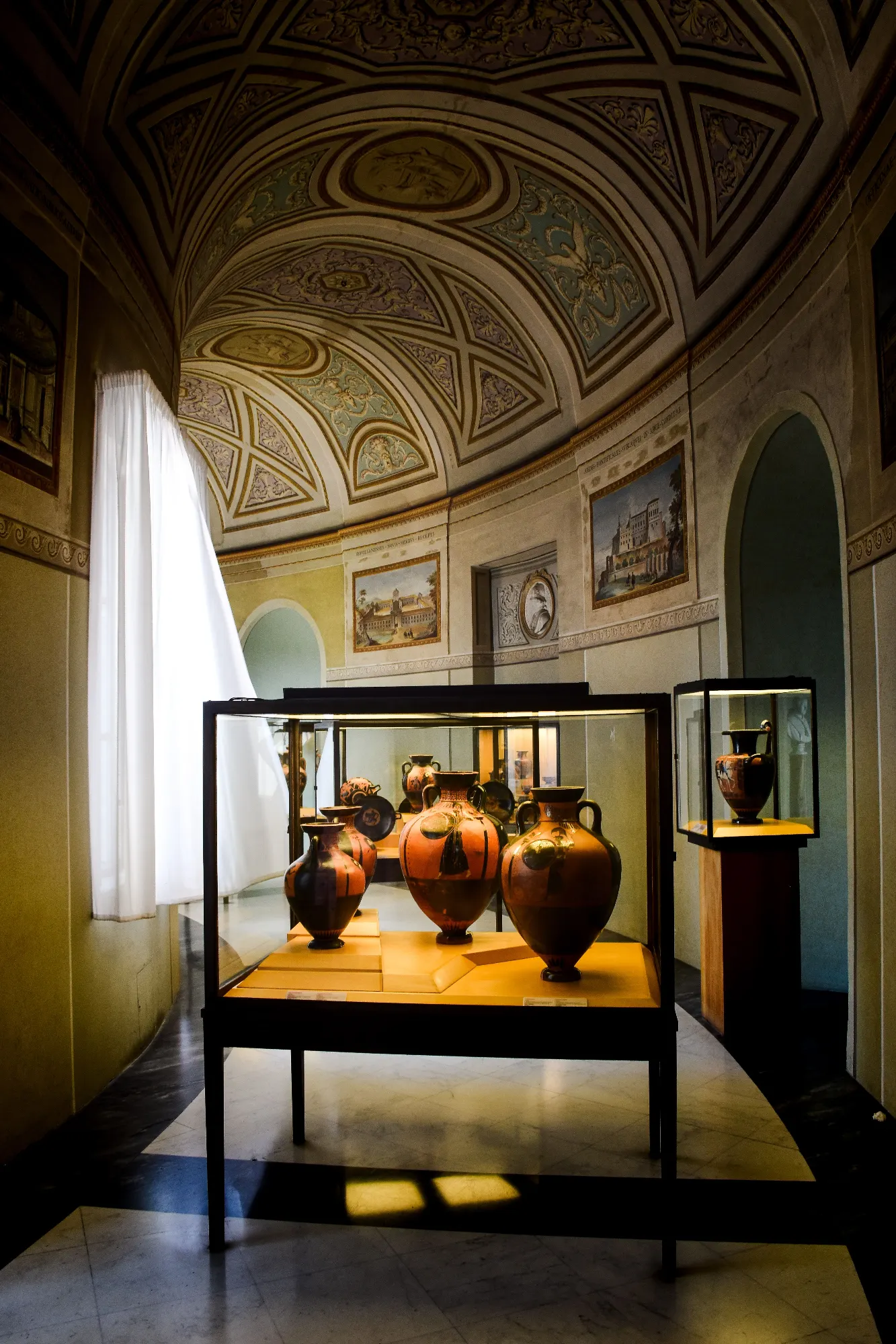A curved museum gallery with a decorated ceiling, displaying ancient Greek vases in glass cases.