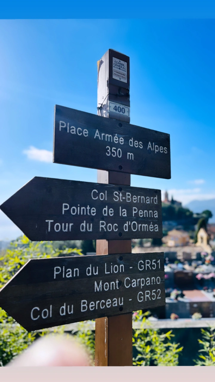 Wooden hiking signpost in Sainte-Agnès showing directions to GR51 and GR52 trails against a blue sky.