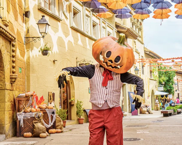 Performer in a pumpkin head and scarecrow costume poses on a decorated street beneath hanging umbrellas.