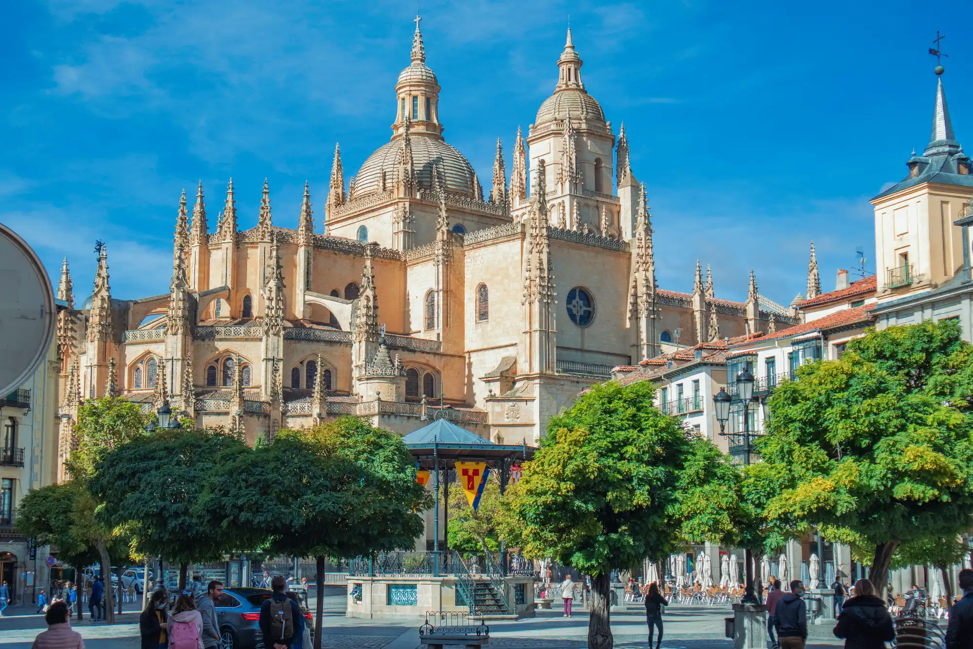 The Gothic Cathedral of Segovia rises above the trees and buildings of the Plaza Mayor on a sunny day.