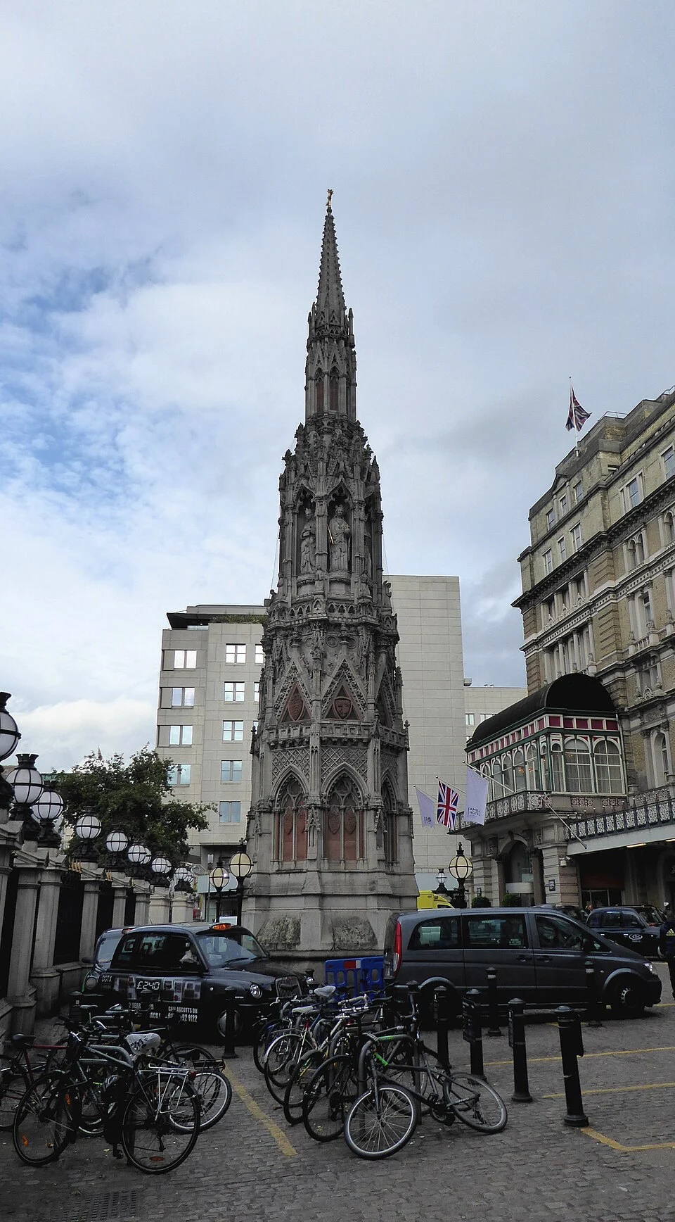 Ozdobny, gotycki pomnik krzyża Eleonory na Charing Cross. Na pierwszym planie widać czarną taksówkę i zaparkowane rowery.