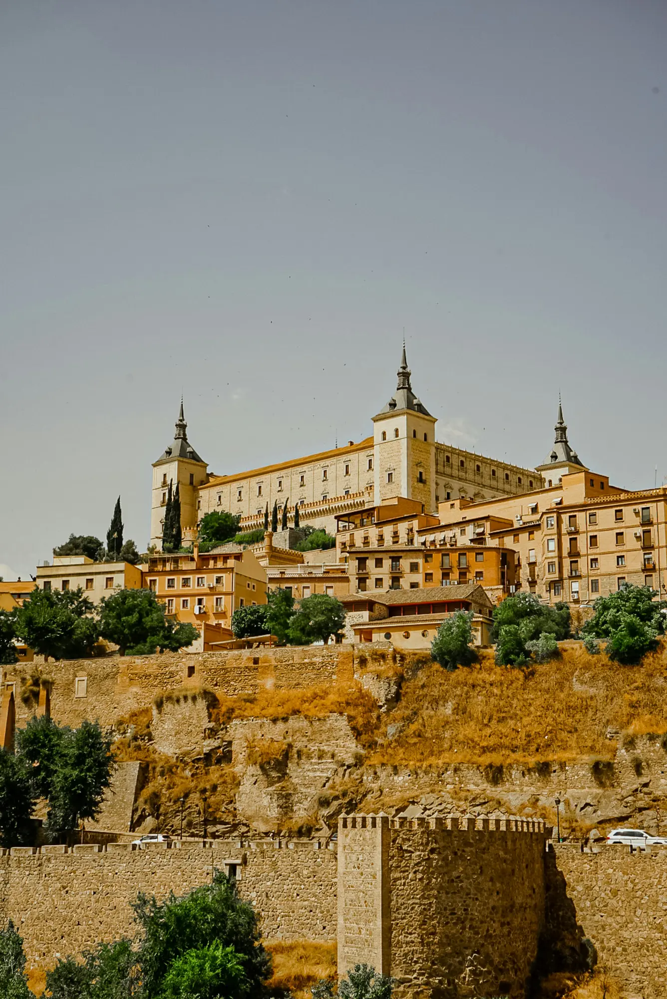 The Alcázar of Toledo rises above the city's stone buildings and defensive walls on a sunny day.