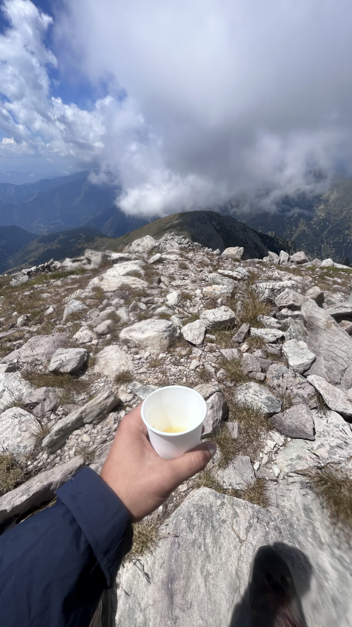Hand holding a small paper cup on a rocky mountain summit with clouds and ridges in the background.