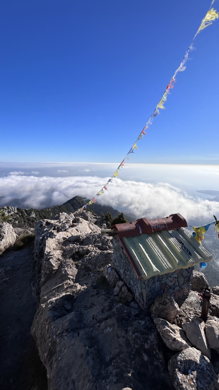 Menton mountain summit with a small stone shrine, prayer flags, and a sea of clouds below under a blue sky.