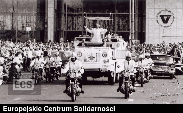 Czarno-białe zdjęcie papieża Jana Pawła II w papamobile, pozdrawiającego tłumy przed Hotelem Victoria w Warszawie.