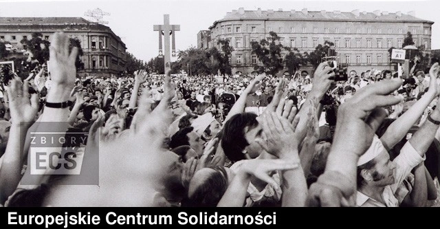 Czarno-białe zdjęcie wielkiego tłumu na placu w Warszawie, z wieloma rękami wzniesionymi do nieba przed dużym krzyżem.