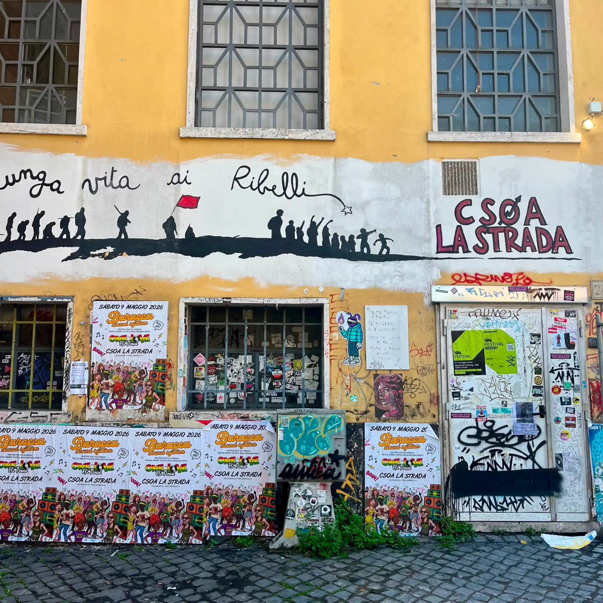 A yellow building facade with a mural, graffiti, and posters for an event at the CSOA La Strada social center.