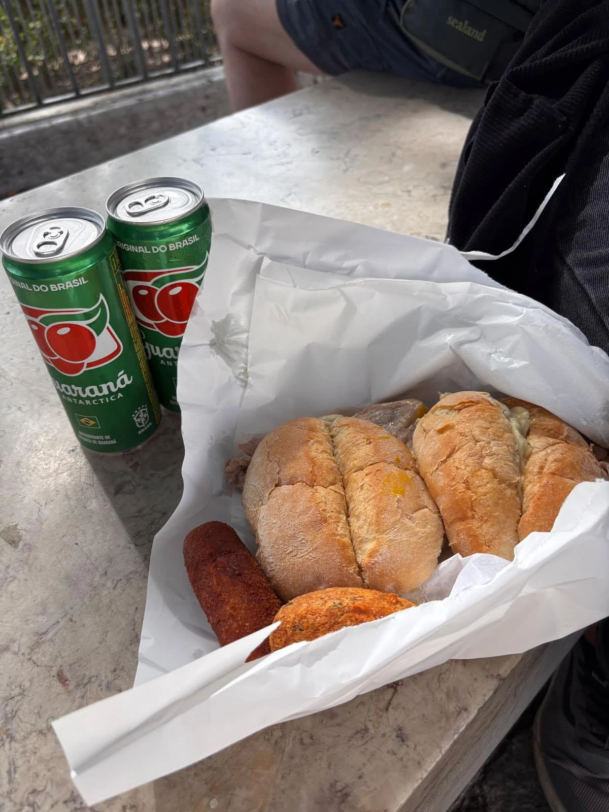 Two pork sandwiches, fried snacks, and two cans of Guaraná soda in a paper bag on a stone bench.