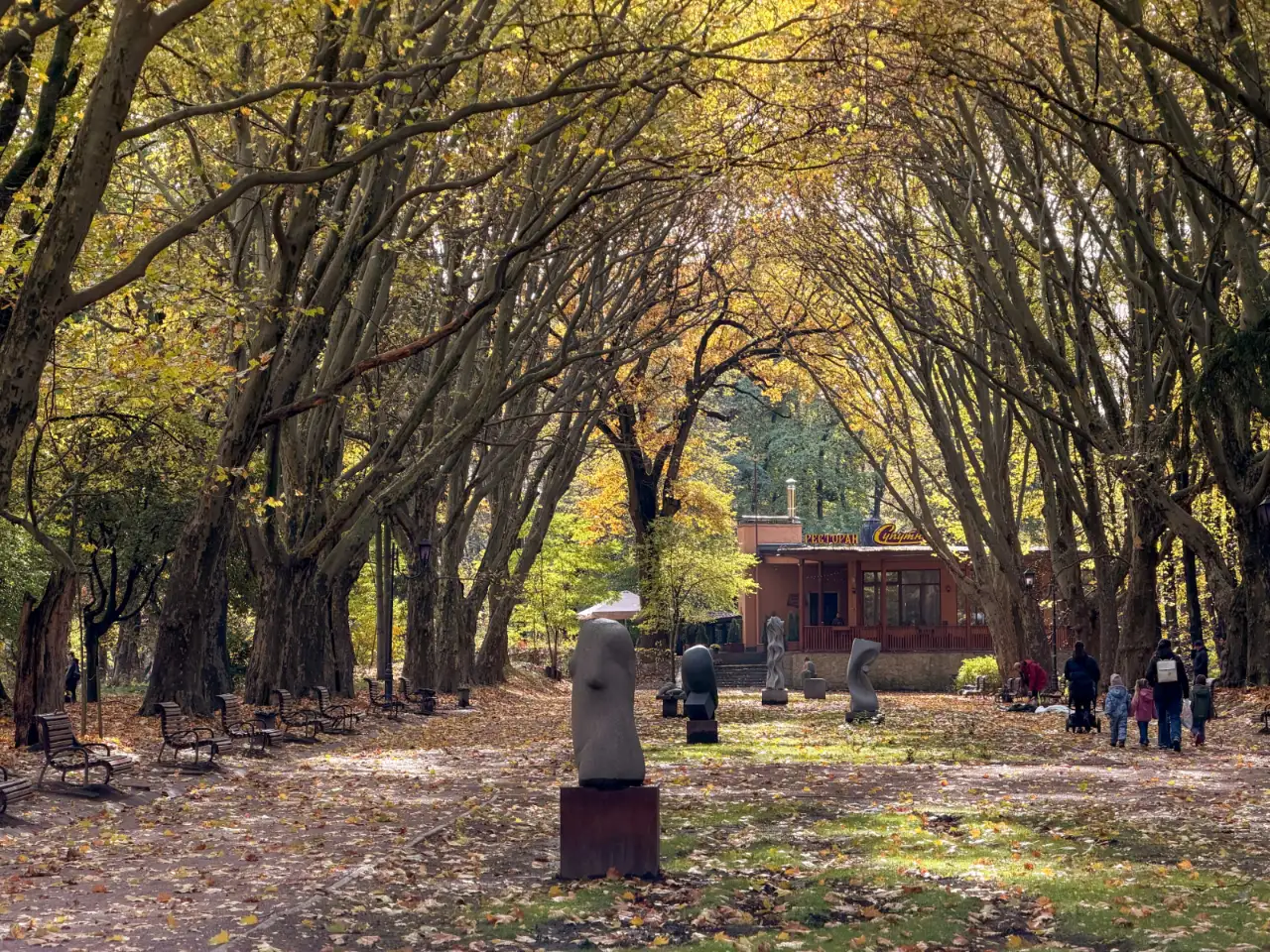 Tree-lined autumn park avenue with modern stone sculptures, benches, and pedestrians.