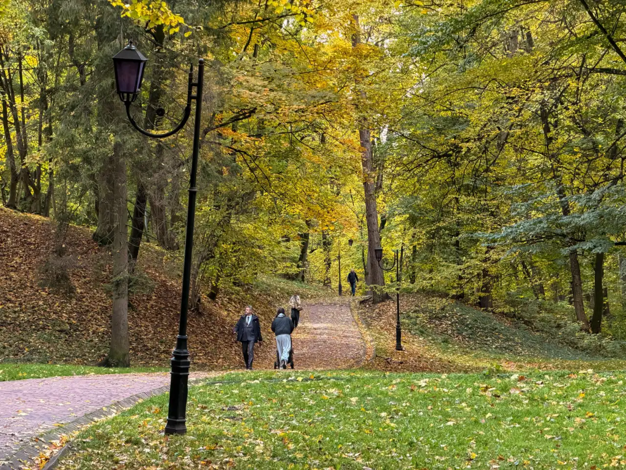 Paved walkway winding through a leafy autumn park with classic lampposts and pedestrians.