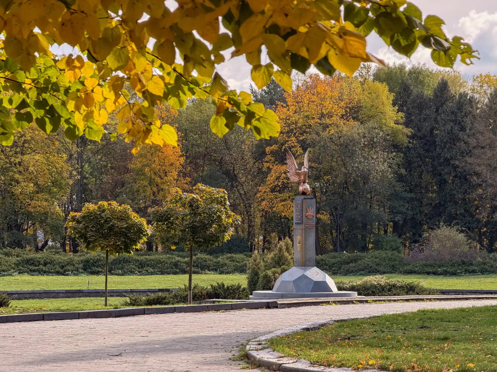 A stone monument with a bird sculpture on a pillar along a park path surrounded by autumn trees and lawns.