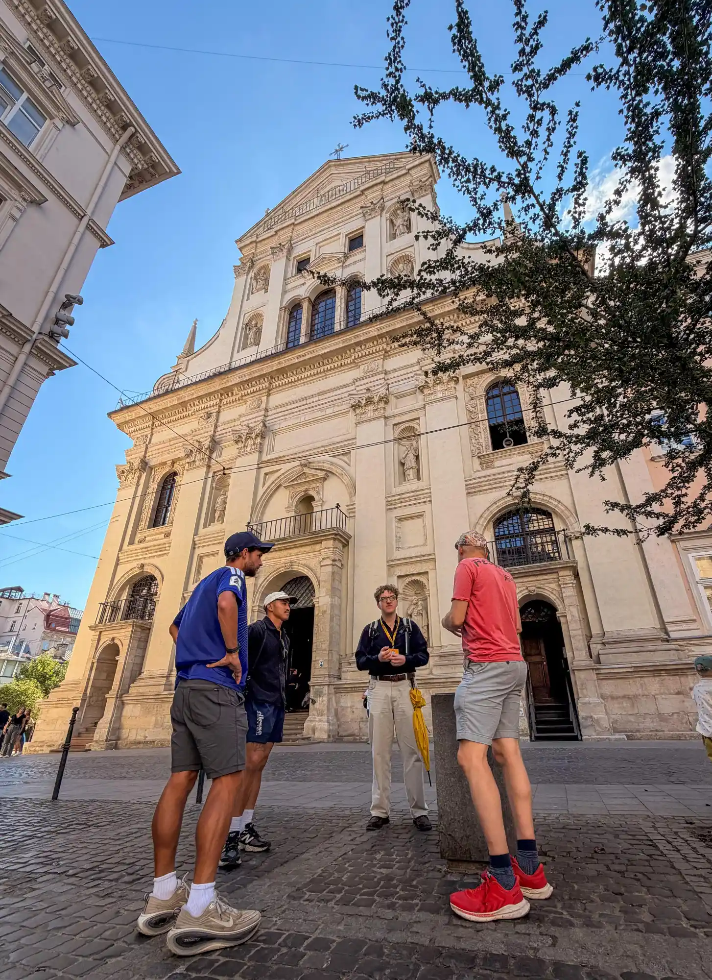 Przewodnik z żółtym parasolem rozmawia z turystami przed Kościołem Jezuitów we Lwowie.