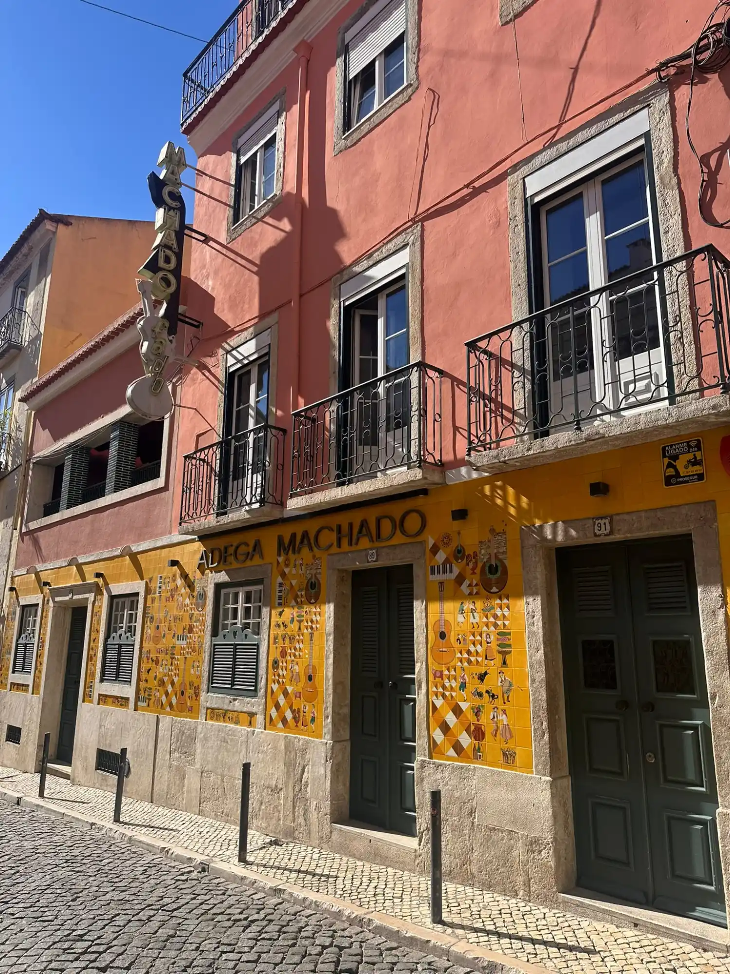 Pink and yellow facade of Adega Machado with decorative tiles and balconies along a stone street in Lisbon.