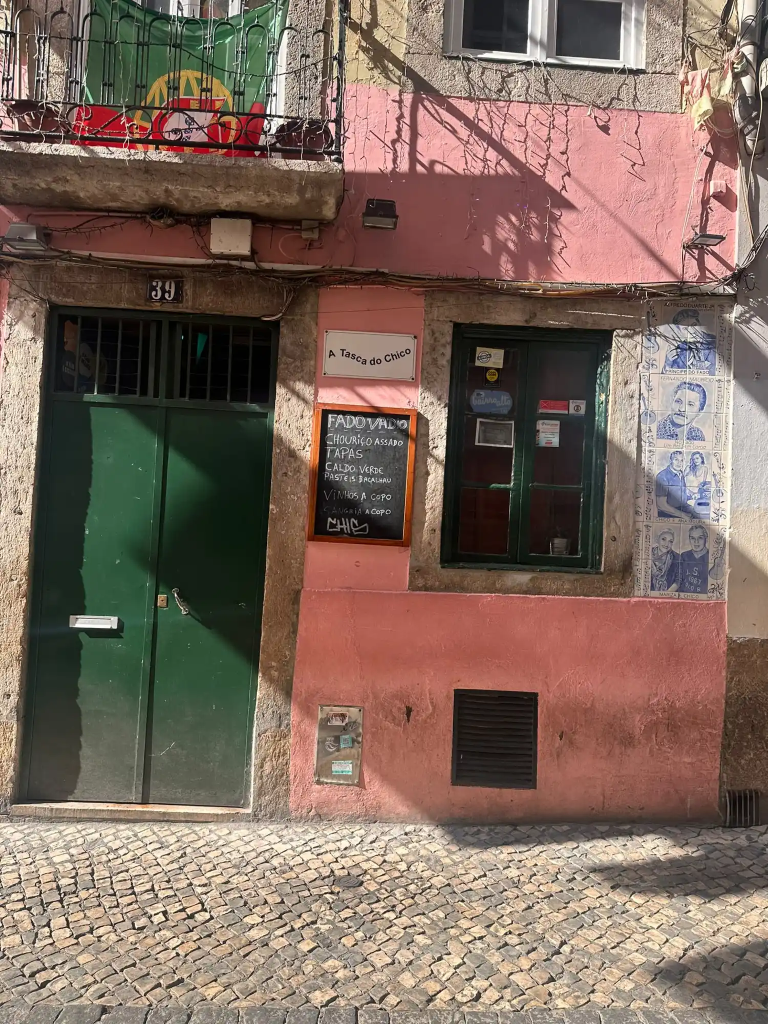 Pink facade of A Tasca do Chico tavern with a green door, chalkboard menu, and cobblestones in Lisbon.