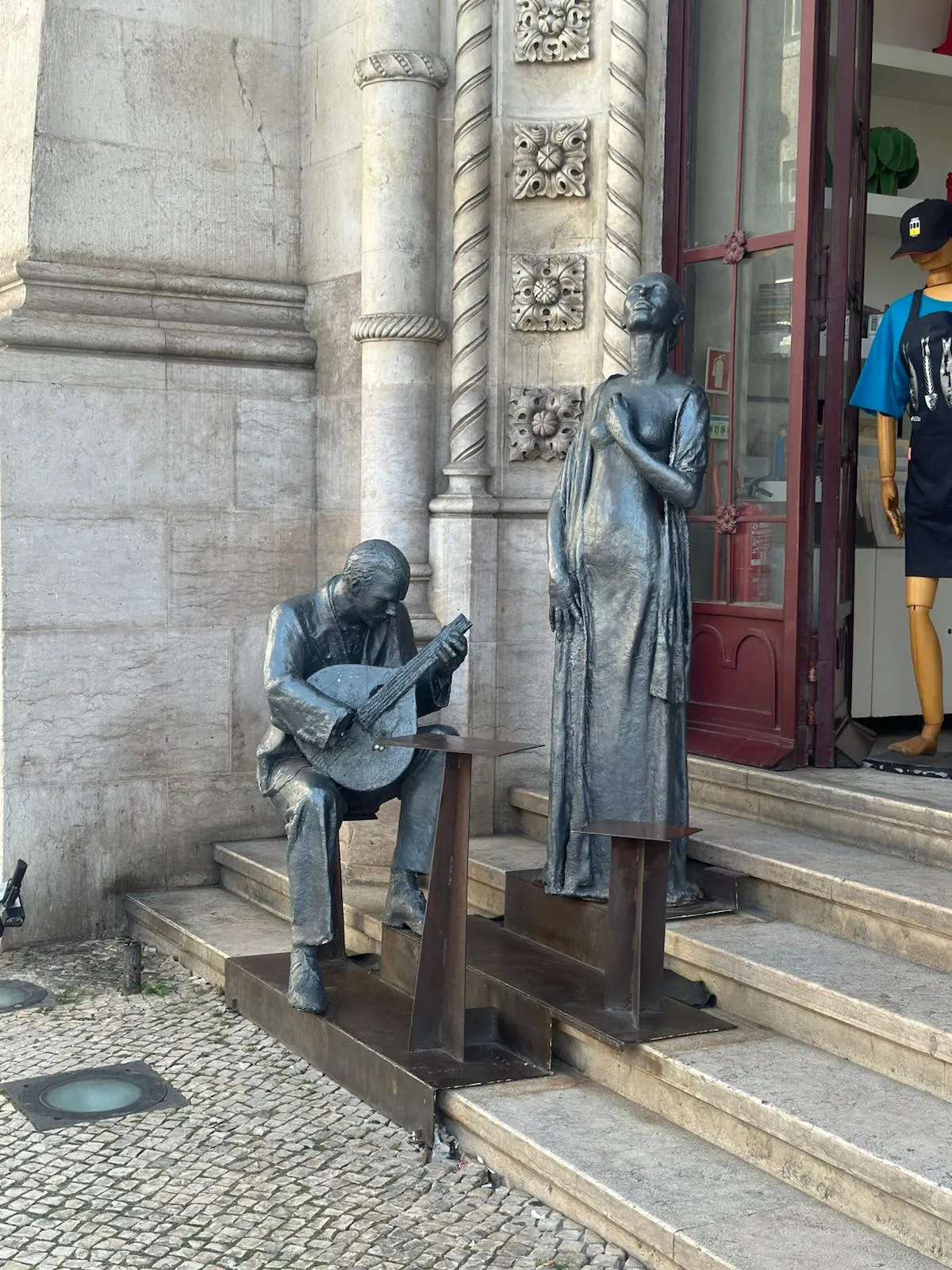 Bronze sculptures of a seated Portuguese guitar player and a standing female fado singer on stone steps in Lisbon.