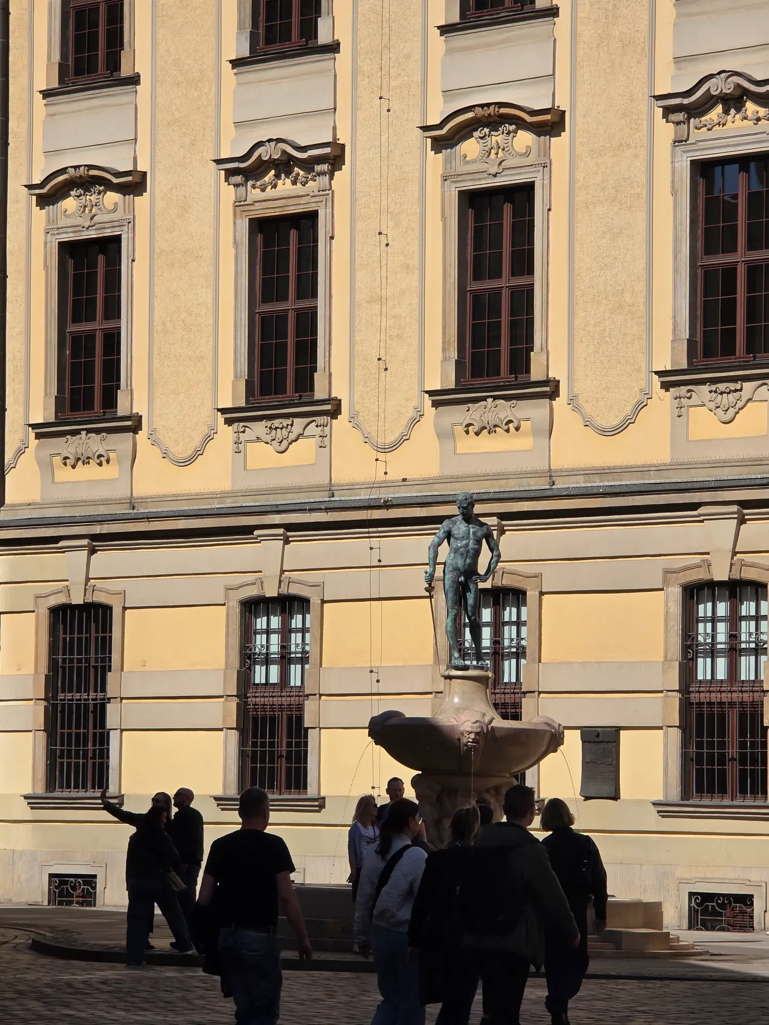 Bronze Fencer Fountain statue in front of the yellow facade of the University of Wroclaw, with tourists walking nearby.