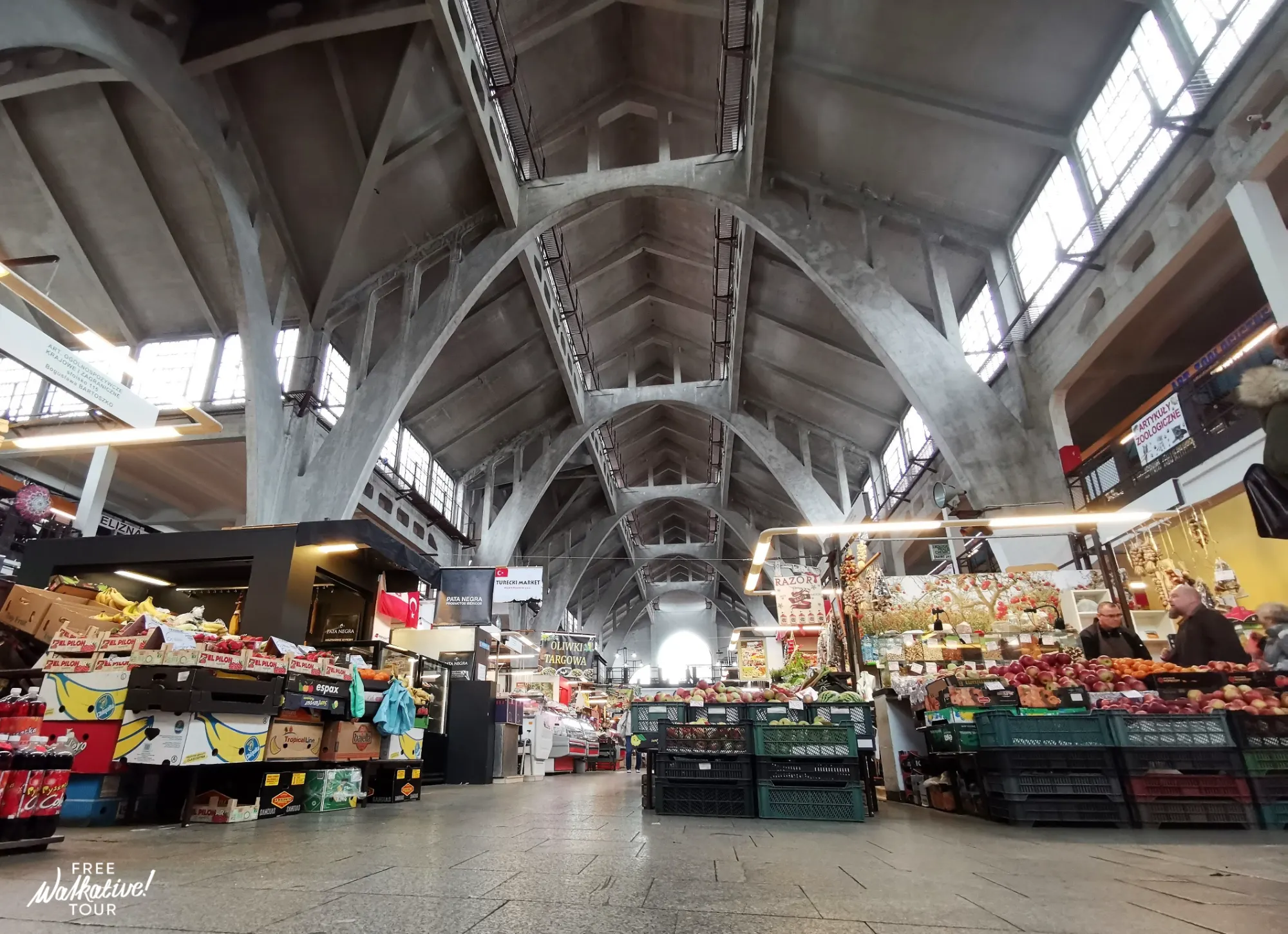 Wnętrze Hali Targowej we Wrocławiu z betonowymi łukami i stoiskami ze świeżymi produktami.