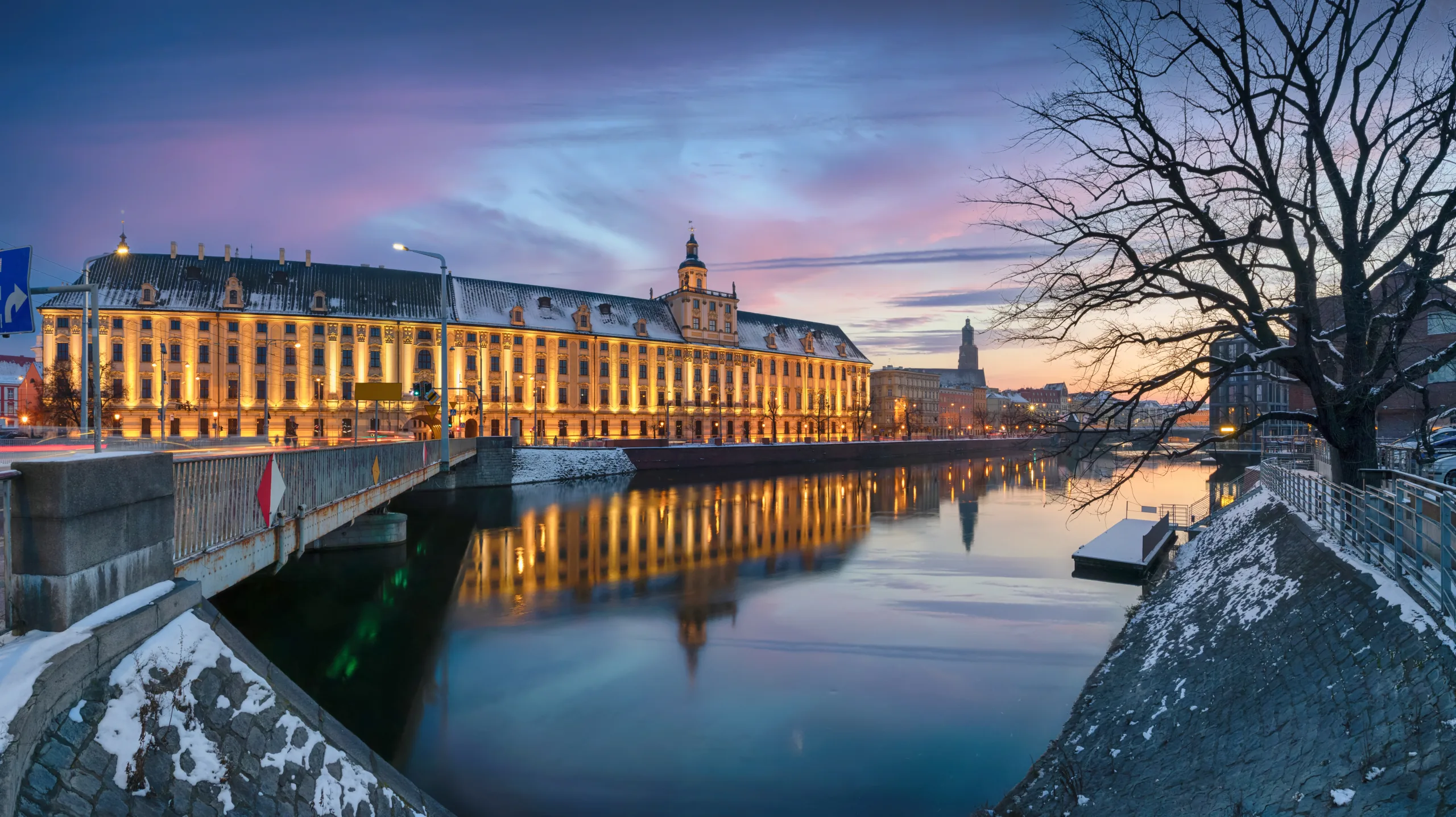 Illuminated University of Wrocław building and bridge over the Odra River at dusk, with snow on banks.
