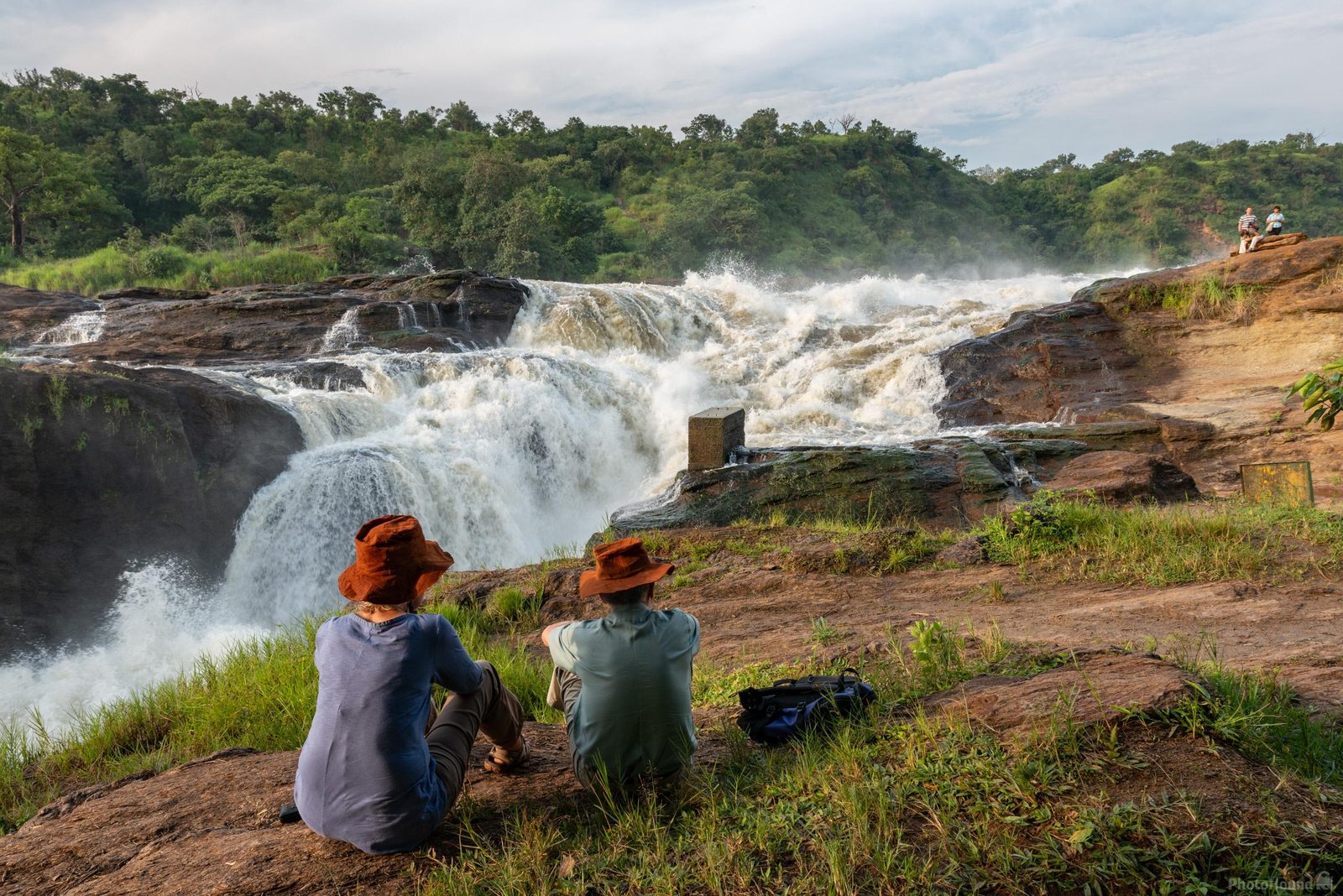 Two people sitting on a rock by a waterfall, surrounded by greenery.
