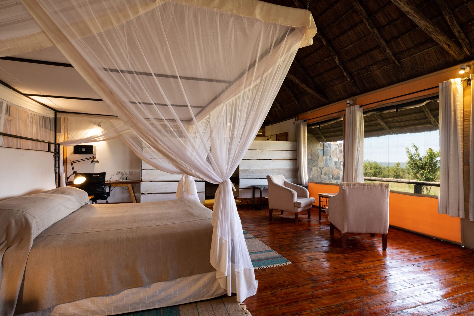 A spacious bedroom with a large bed, a writing desk, and seating area, featuring a thatched roof and wooden flooring.