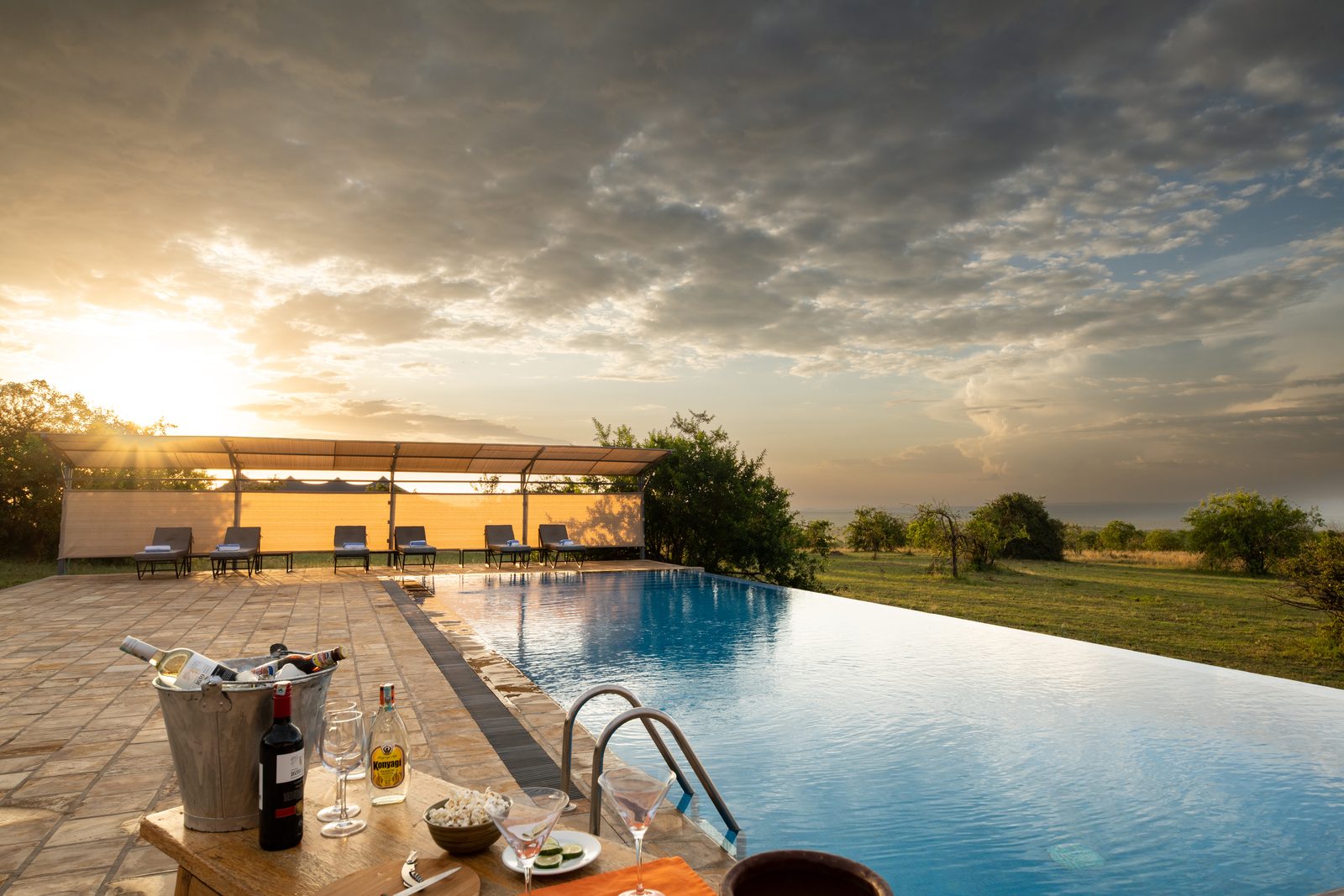 A scenic view of a swimming pool at sunset, with lounge chairs and a table featuring drinks and snacks.