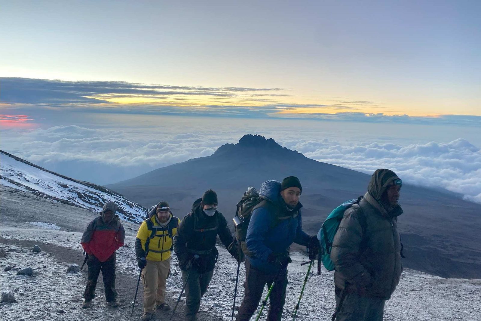 A group of hikers ascending a mountain with a view of clouds and a peak in the background during sunrise.