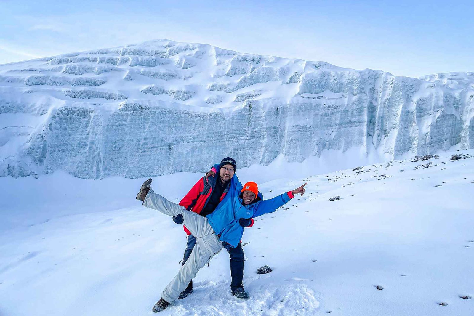 Two people posing playfully in front of a large ice formation on a snowy landscape.