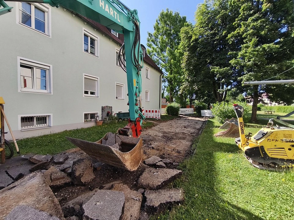 Bagger der Firma Hartl mit Schaufel auf aufgebrochenem Asphaltweg neben einem Wohnhaus in der Franz-Xaver-Bauer-Straße, rechts eine Rüttelplatte auf der Wiese